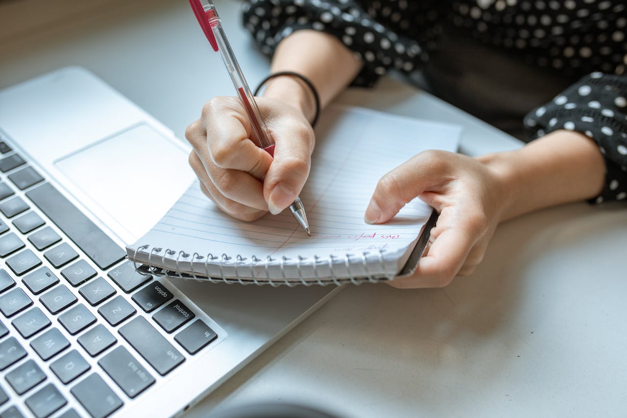 Person writing in a spiral notebook next to a laptop, capturing a planning moment.